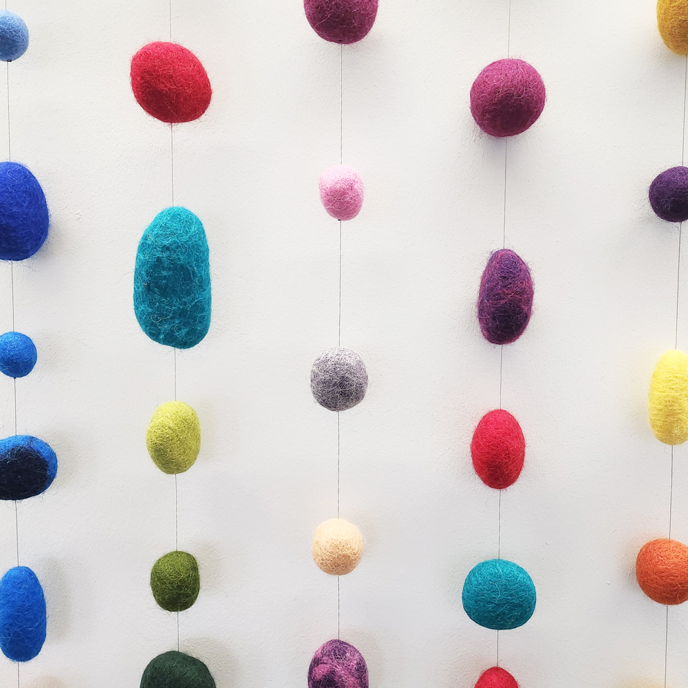 Sjónlýsing: Litaðir, filtaðir ullarhnútar í ýmsum stærðum hanga á þunnum þráðum í lóðréttum röðum á hvítum vegg. Áferðin er loðin og mjúk, með fíngerðum trefjum sem sjást í nærmynd. Formin hanga með reglulegu bili og mynda taktfast mynstur; daufir skuggar undir hverju formi gefa dýpt og tilfinningu fyrir léttleika og hreyfingu.